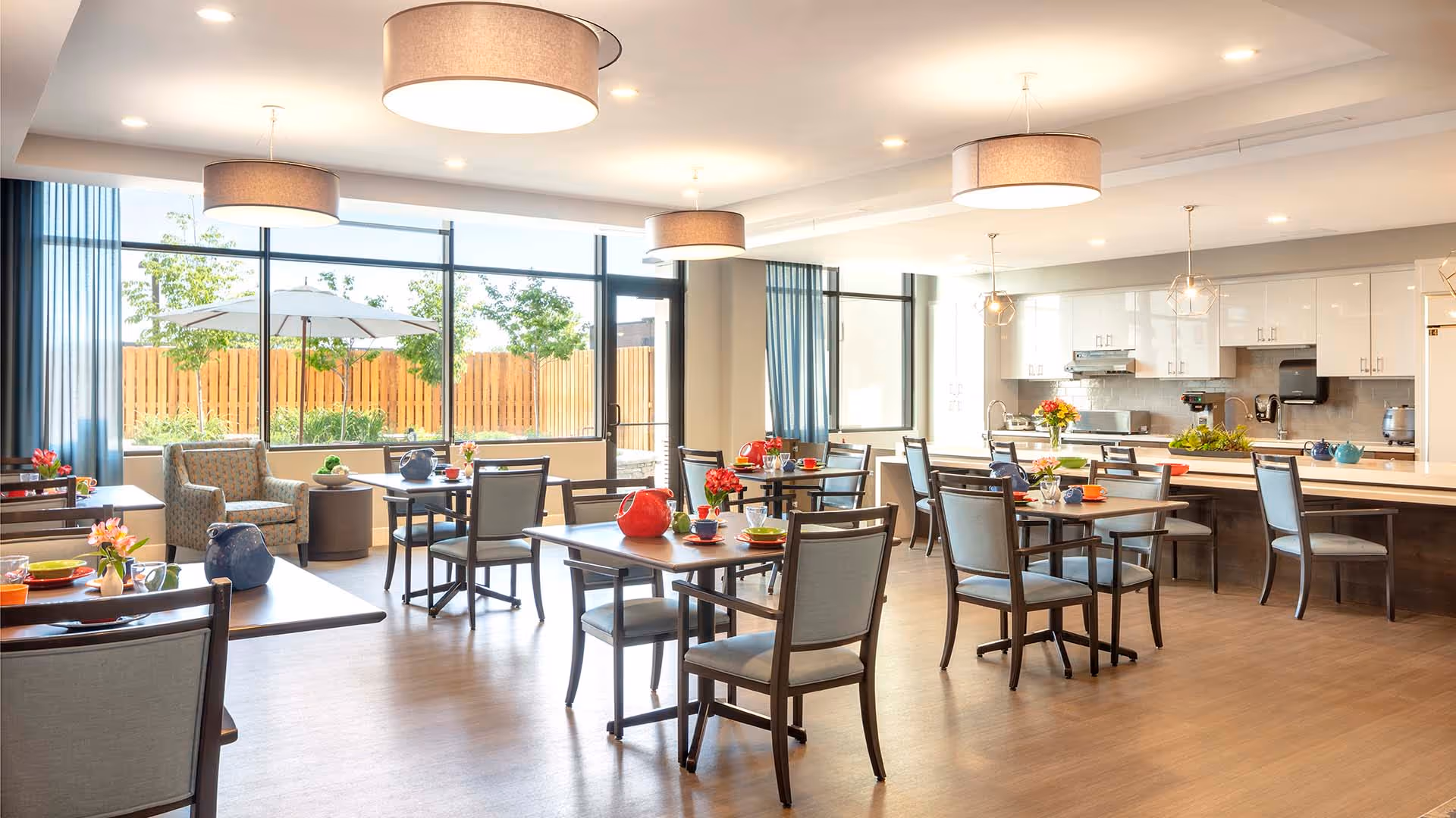 A bright and spacious dining room in a senior living facility with multiple tables and chairs arranged neatly. The room features large windows letting in natural light, modern pendant lights, and a kitchen area with white cabinets and countertops in the background. There are colorful teapots and flowers on the tables, and a cozy seating area with armchairs near the windows.