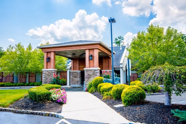 Entrance to a building with a covered driveway supported by stone and wood pillars, surrounded by well-maintained landscaping including green bushes, flowering plants, and trees under a partly cloudy sky.