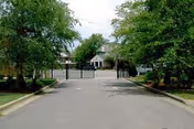 View of a gated entrance to a residential facility with a paved driveway, surrounded by green trees and bushes on both sides, leading to a building in the background.