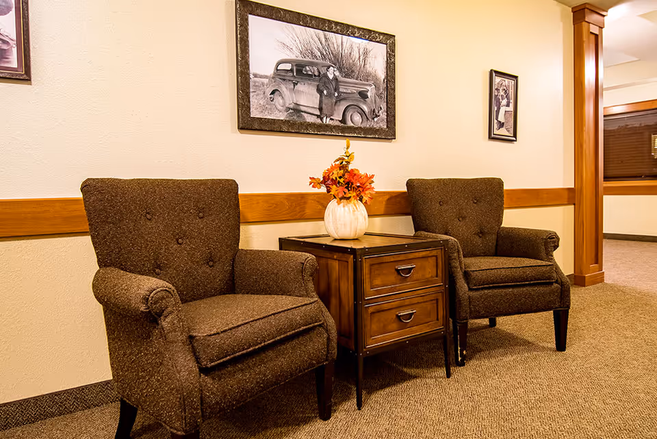 Two brown upholstered armchairs with button tufting are placed on either side of a wooden side table with two drawers. On the table is a white pumpkin vase with autumn-colored flowers. The wall behind features a wooden chair rail and two framed black and white vintage photographs. The carpeted floor and warm lighting create a cozy atmosphere.