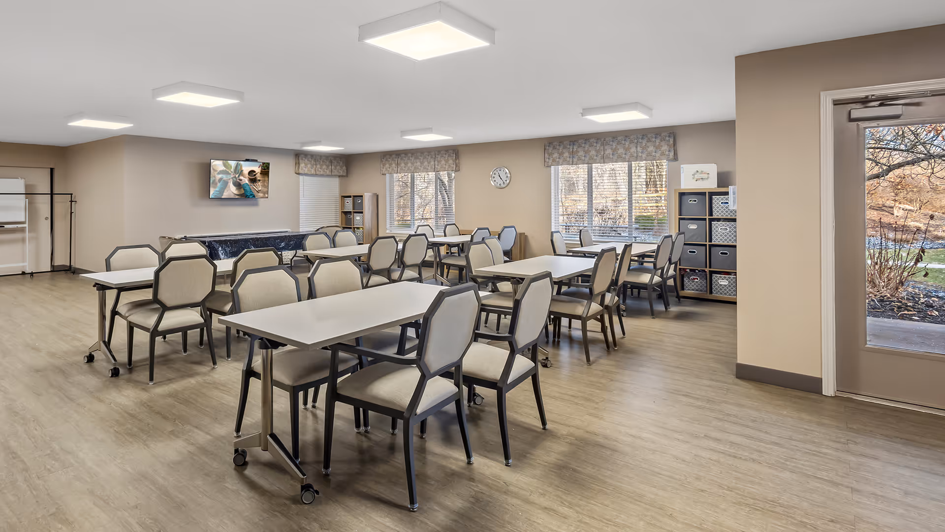 A bright and spacious dining room with several tables and cushioned chairs arranged neatly. The room has large windows with floral valances letting in natural light, a wall clock, and storage shelves with bins. A door leads outside to a garden area visible through the glass. The floor is light wood, and the walls are painted beige.