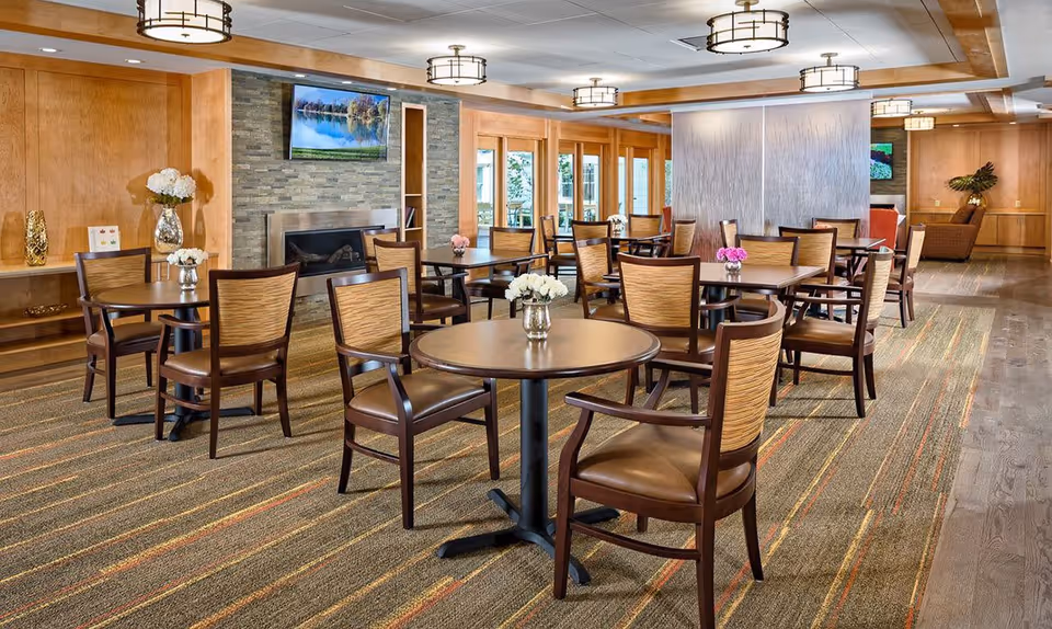 A spacious dining room with multiple round and square tables surrounded by wooden chairs with cushioned seats. The room features a carpeted floor with a striped pattern, a stone fireplace with a mounted TV above it, and large windows allowing natural light. There are decorative flowers on the tables and modern ceiling lights.