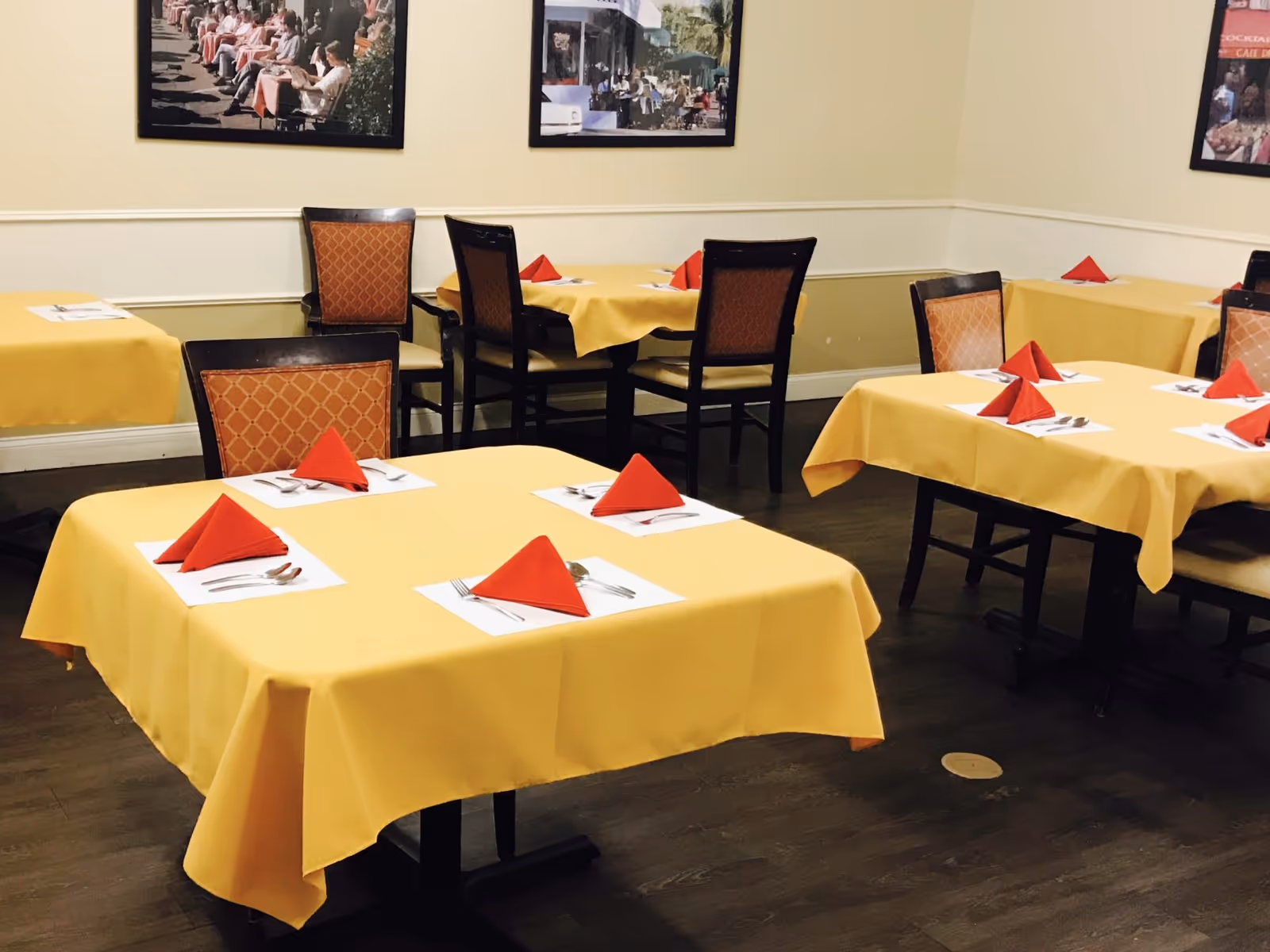 Dining room with several tables covered in yellow tablecloths, each set with white placemats, silverware, and red folded napkins. The room has wooden chairs with orange patterned cushions and framed pictures on the beige walls.