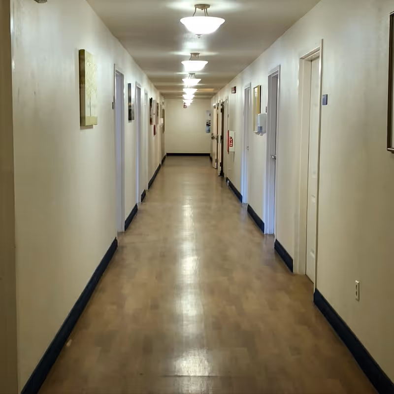 Long, well-lit indoor hallway with multiple closed doors and framed artwork along the walls.
