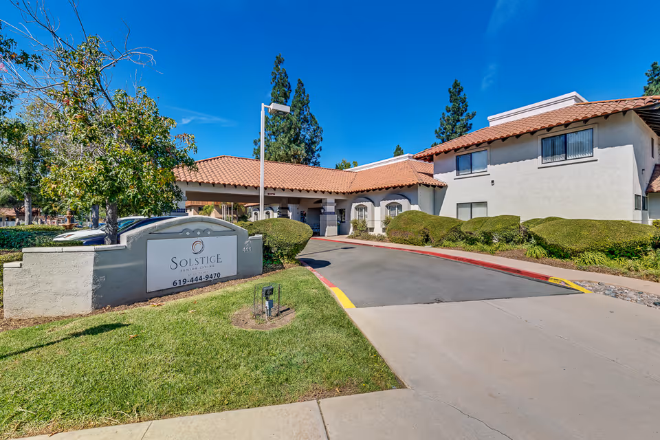 Exterior front entrance of Solstice Senior Living with driveway, entry canopy, landscaped shrubs and a sign in the foreground.