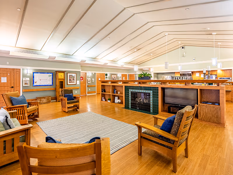 A spacious and well-lit common area with wooden furniture including chairs and sofas arranged around a striped area rug. The room features a central fireplace with green tiles and built-in wooden shelving. The ceiling is vaulted with recessed lighting and hanging pendant lights. The walls are decorated with framed pictures and a bulletin board, and there are multiple wooden doors leading to other rooms.