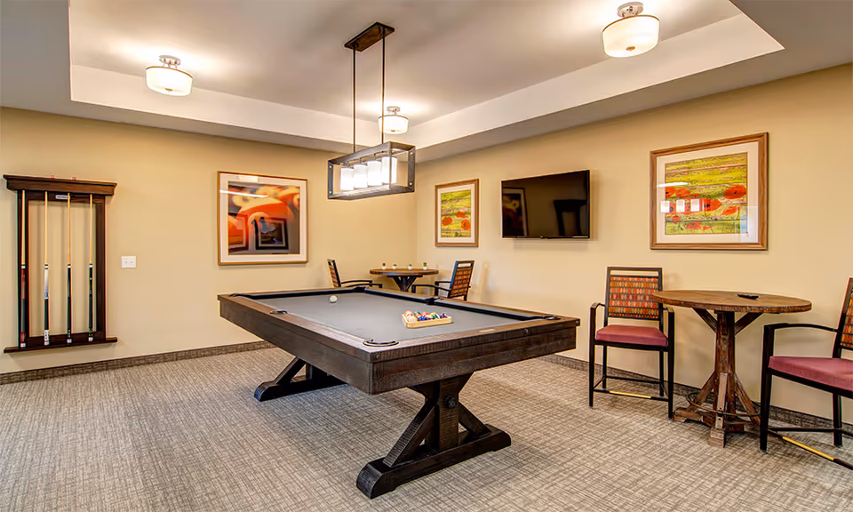 A recreational room with a pool table in the center, surrounded by chairs and small round tables. The walls are decorated with framed artwork, and a flat-screen TV is mounted on one wall. The room has carpeted flooring and ceiling lights.