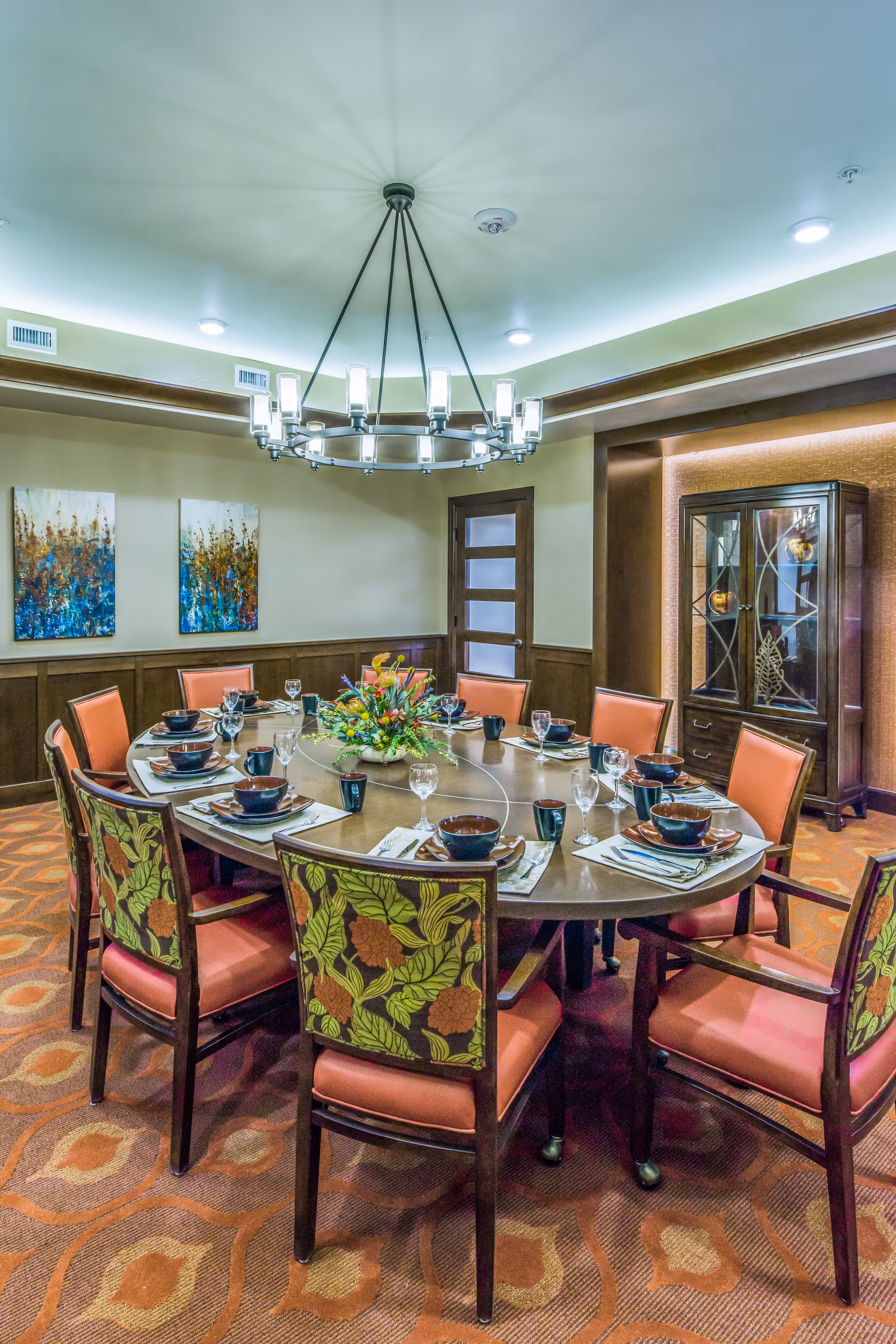 A well-lit dining room with a large oval wooden table set for ten people. The chairs have orange cushions and floral patterned backs. The table is set with bowls, plates, glasses, and mugs, and a floral centerpiece is in the middle. The room features a modern chandelier, two abstract paintings on the wall, a wooden cabinet with glass doors, and a patterned carpet.