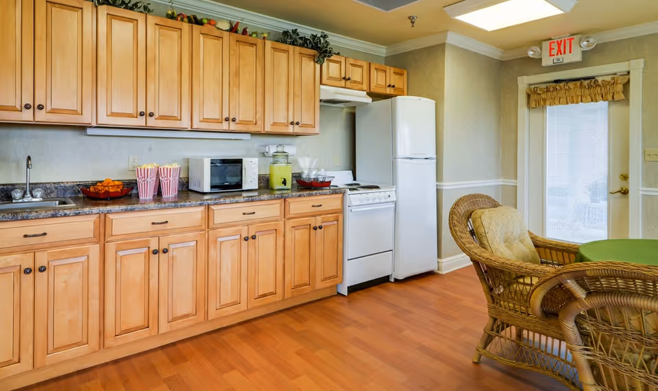 Small communal kitchen with light wood cabinets, countertop appliances, a refrigerator and stove, and a wicker dining table and chairs by an exit door.