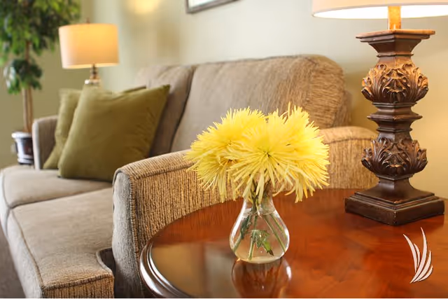 A cozy living room area featuring a beige textured sofa with green cushions, a wooden side table with a clear vase holding yellow flowers, and an ornate table lamp with a beige shade. A green plant and another lamp are visible in the background.