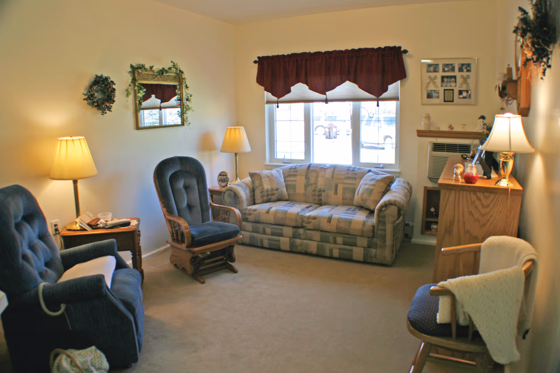 Cozy living room with a patterned sofa under a window, two upholstered chairs, lamps, and wooden furniture.