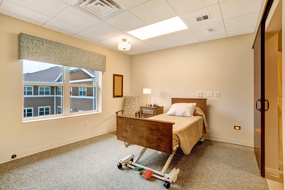 A simple, clean bedroom in a senior living facility with a single hospital-style bed, a wooden nightstand with a lamp and phone, a patterned armchair, a large window with a view of neighboring buildings, and a sliding door closet.