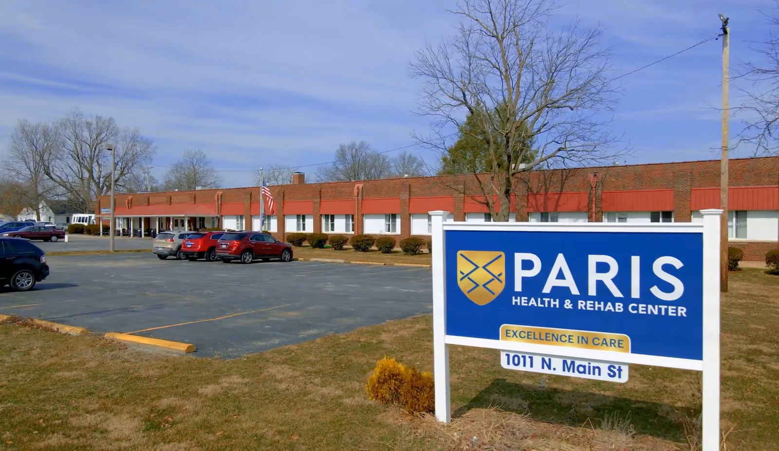 Exterior view of Paris Health & Rehab Center, a single-story brick building with a red roof trim. Several cars are parked in the parking lot in front of the building. A large sign in the foreground displays the facility's name, slogan 'Excellence in Care,' and address '1011 N. Main St'. Leafless trees and a clear sky are visible in the background.