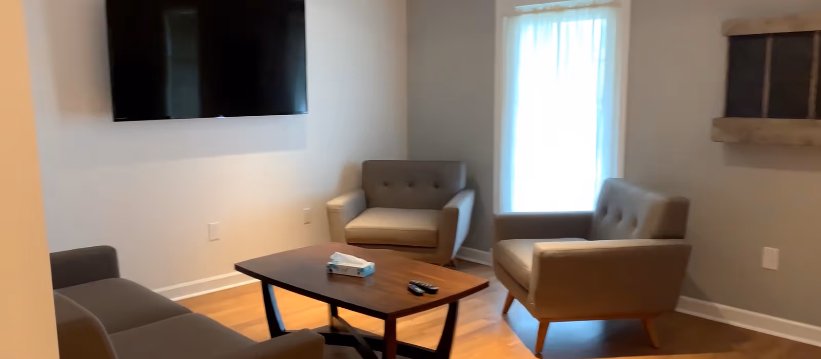 Small sitting area with three mid-century armchairs around a coffee table, a wall-mounted TV, and a window with a sheer curtain.