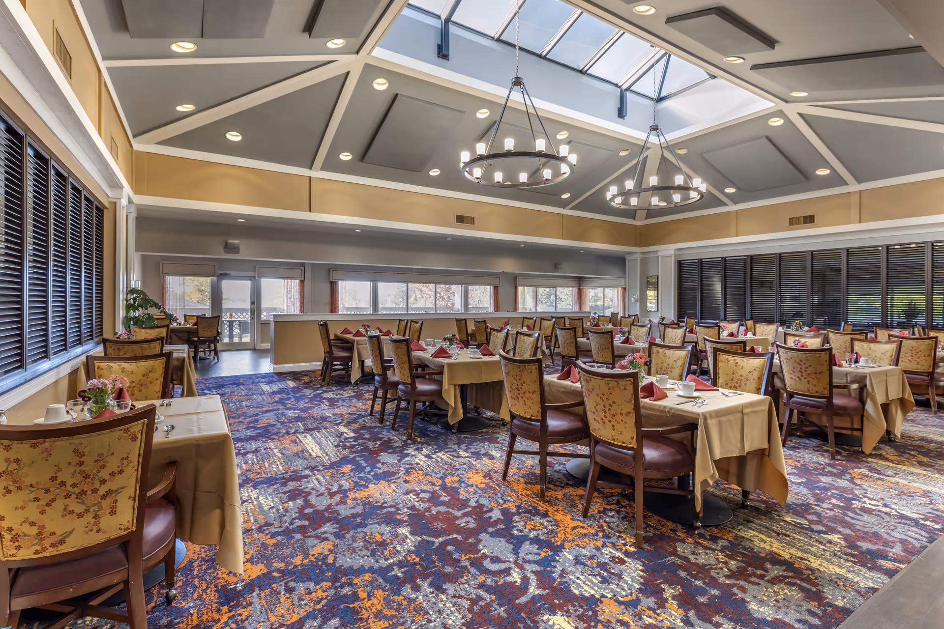 A spacious dining room with multiple tables covered in beige tablecloths, each set with cups, utensils, and red folded napkins. The chairs have floral upholstery and wooden frames. The room features a colorful patterned carpet in shades of blue, orange, and gray. Large windows and glass doors allow natural light to enter, and two circular chandeliers hang from a high ceiling with skylights.
