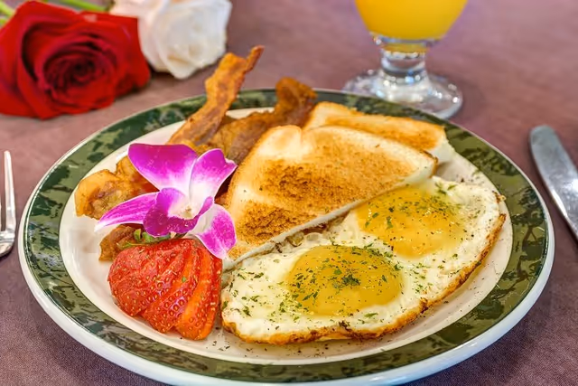 Plate with two sunny-side-up eggs, toast, bacon, a sliced strawberry and a purple orchid garnish on a table with a glass of orange juice and roses in the background.