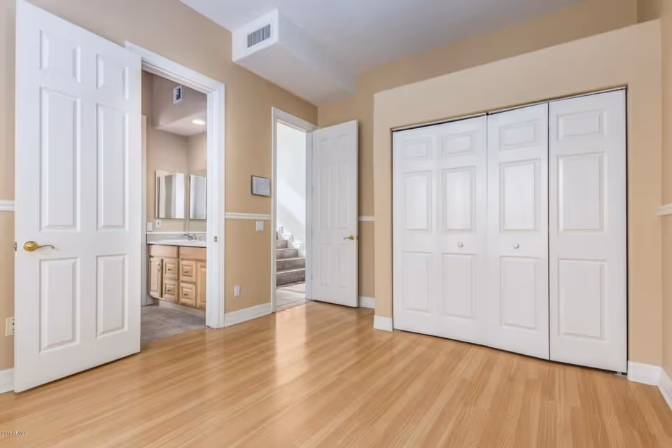 Empty room with light wood flooring and beige walls, featuring two white doors on the left side, one leading to a bathroom with a sink and mirror visible, and the other leading to a hallway with stairs. On the right side, there is a large white closet with double doors.