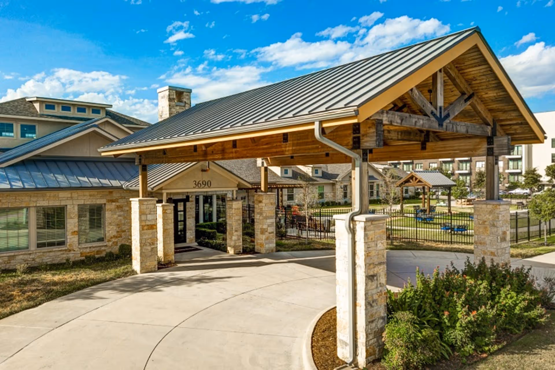 Exterior view of The Auberge at Plano senior living facility showing a covered entrance with stone pillars and a metal roof, a driveway, landscaped greenery, and a clear blue sky with some clouds.
