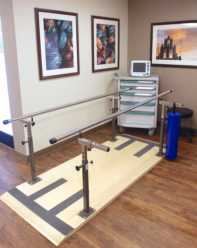 A rehabilitation room with parallel walking bars mounted on a wooden platform, a medical cart, and framed artwork on the walls.