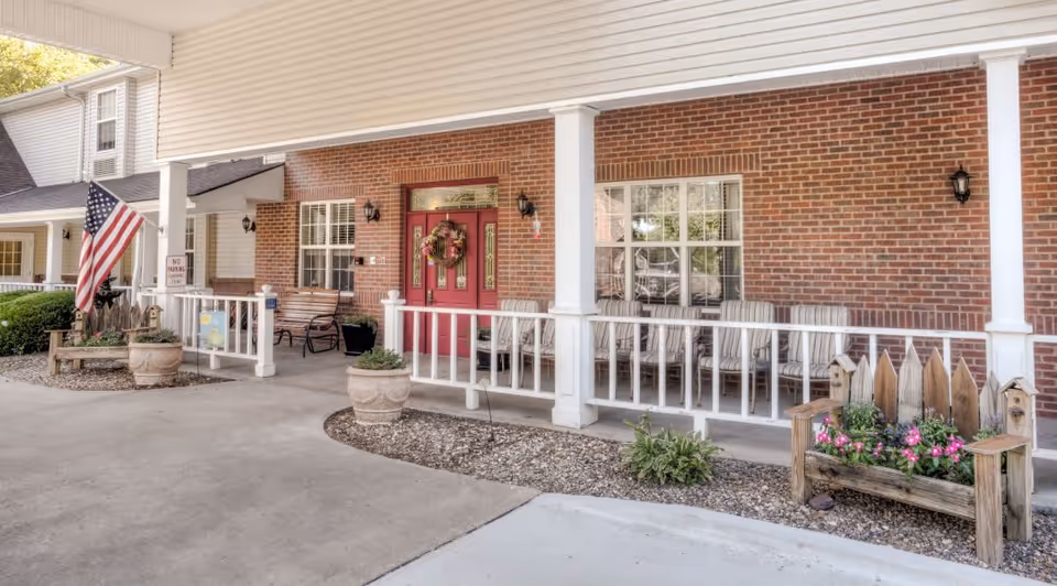 Covered front porch of a brick building with a red door decorated with a wreath, several striped cushioned chairs lined up along the porch, potted plants, an American flag, and a wooden planter with pink flowers.