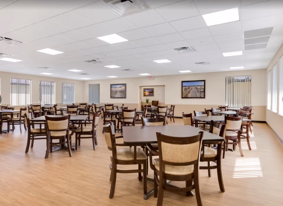 A spacious dining room with multiple dark wood tables and chairs with beige cushions. The room has light-colored walls, several windows with vertical blinds, and framed landscape pictures on the walls. The floor is a light wood laminate, and the ceiling has recessed lighting panels.