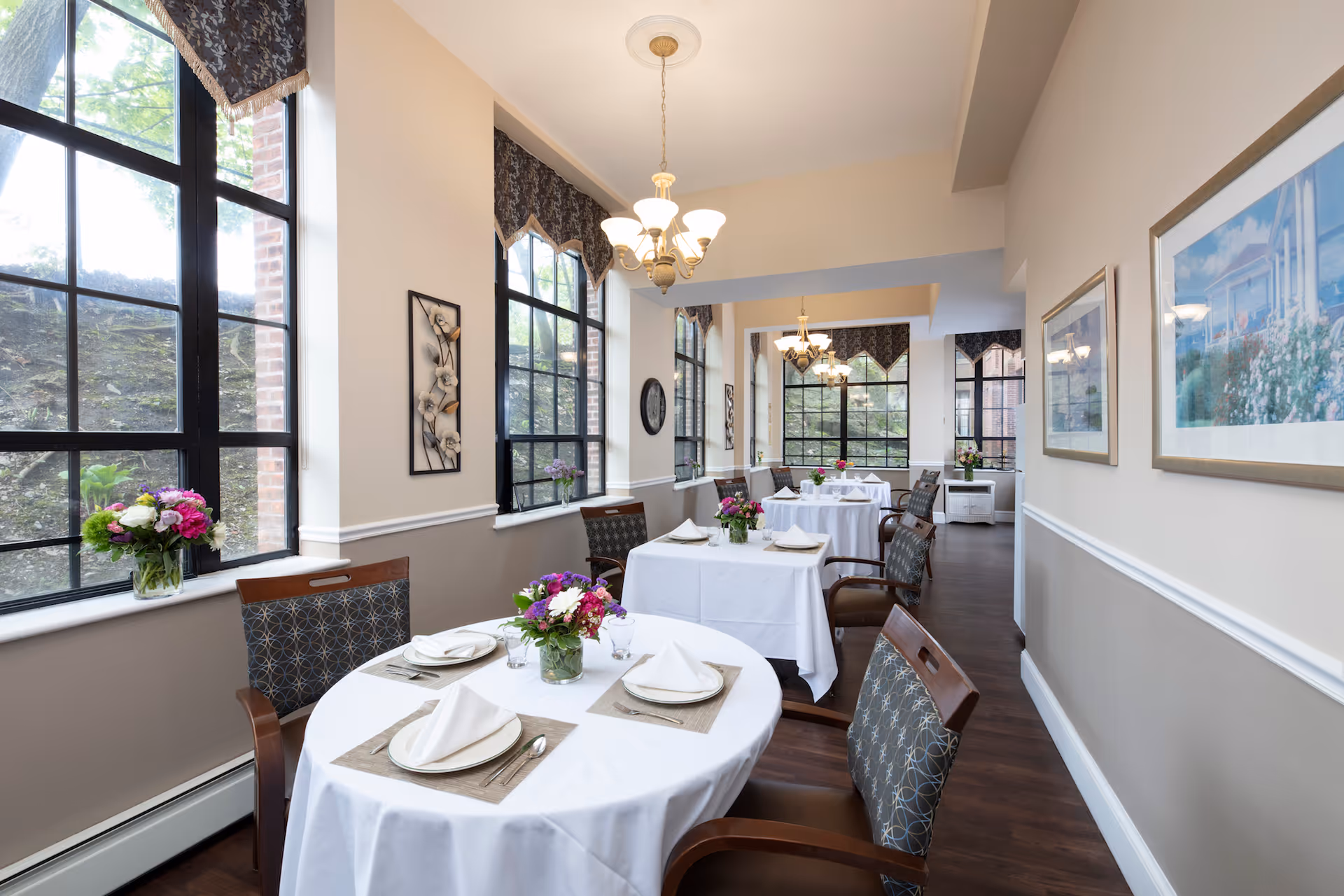 A bright dining room with round tables set with white linens and floral centerpieces, large windows, and chandeliers.
