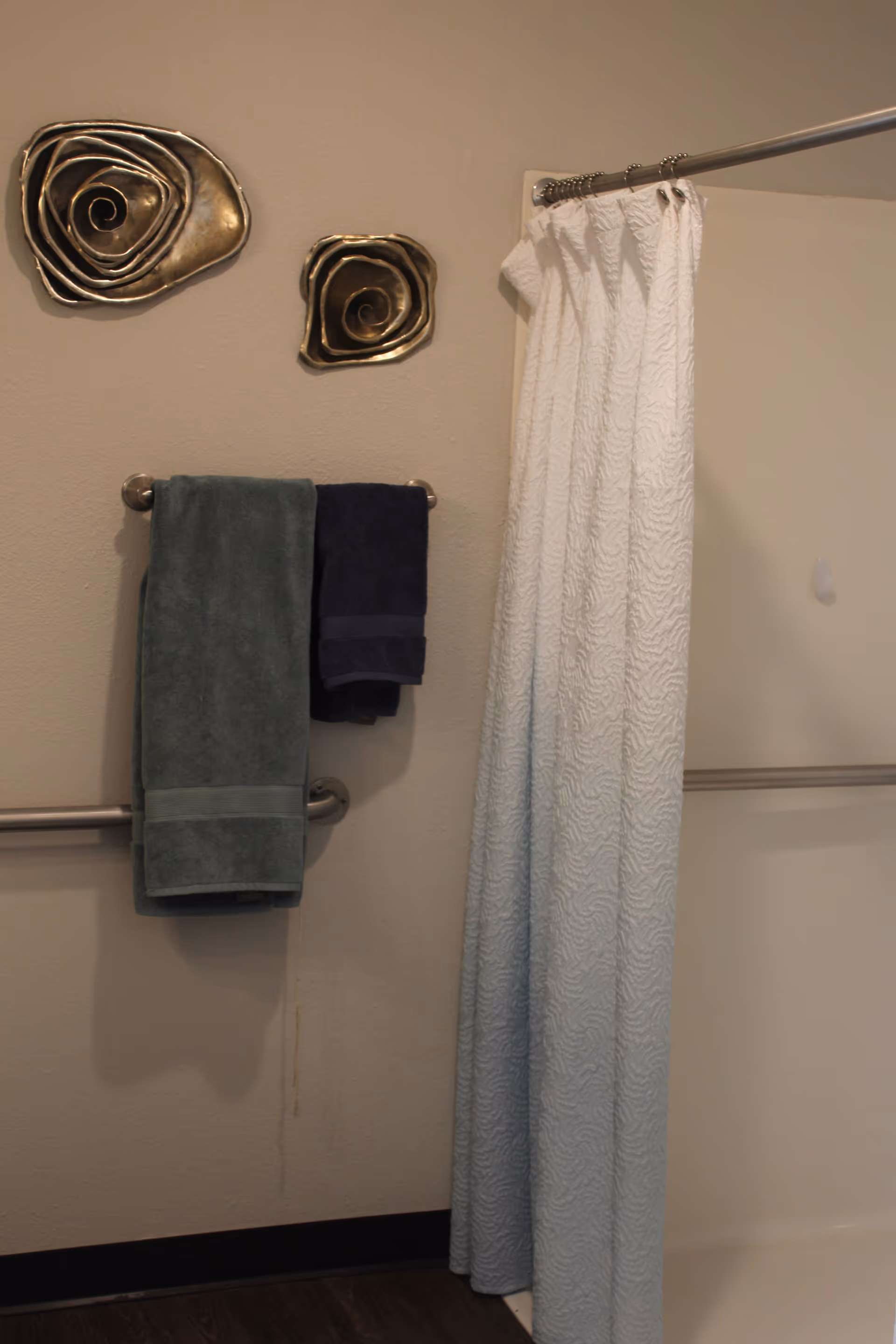 Bathroom interior with a shower and white patterned curtain, a towel rack holding folded towels, and decorative metal wall art.