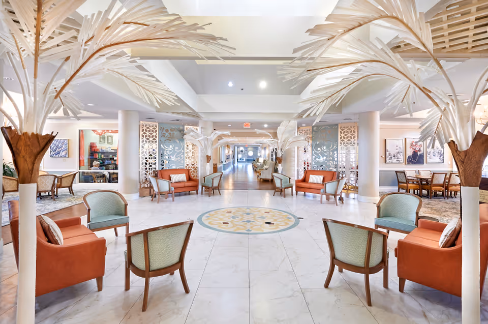 Bright and spacious senior living facility common area with white marble flooring, decorative palm-like sculptures, orange and green cushioned chairs arranged in small seating groups, and a long hallway leading to more seating and dining areas in the background.