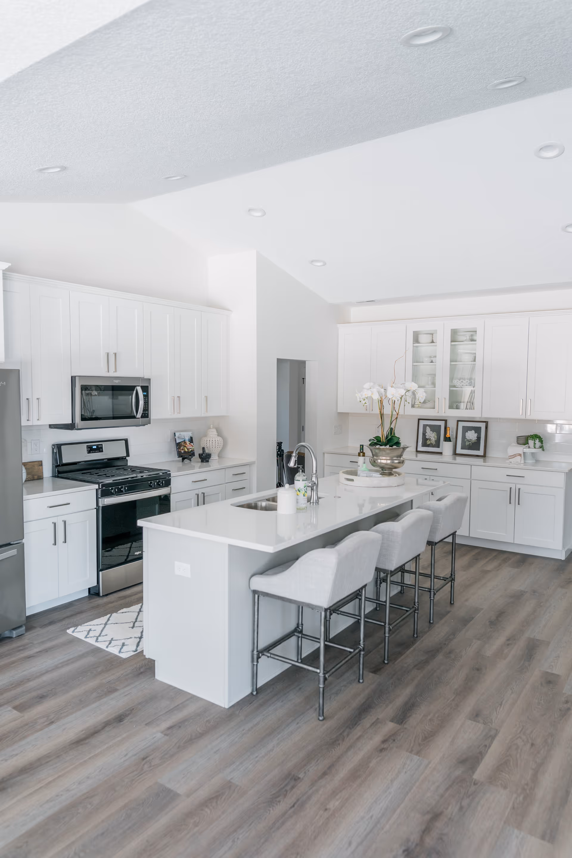 Bright modern kitchen with white cabinetry, stainless steel appliances including a refrigerator, stove, and microwave. A large white island with a sink and faucet has four cushioned bar stools. The kitchen has wood flooring and recessed lighting in the ceiling. Decorative items and framed pictures are placed on the countertops.