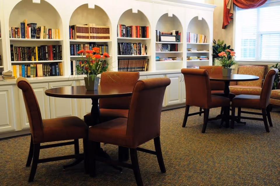 A cozy reading or sitting area with two round tables, each surrounded by four upholstered chairs. Each table has a vase with orange flowers. Behind the tables is a white built-in bookshelf filled with books. A window with white blinds and red curtains lets in natural light, and there are some green plants near the window.