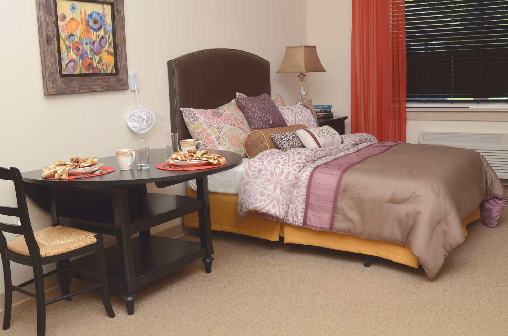 A cozy senior living bedroom with a made bed, decorative pillows, bedside lamp and a small table set for two.
