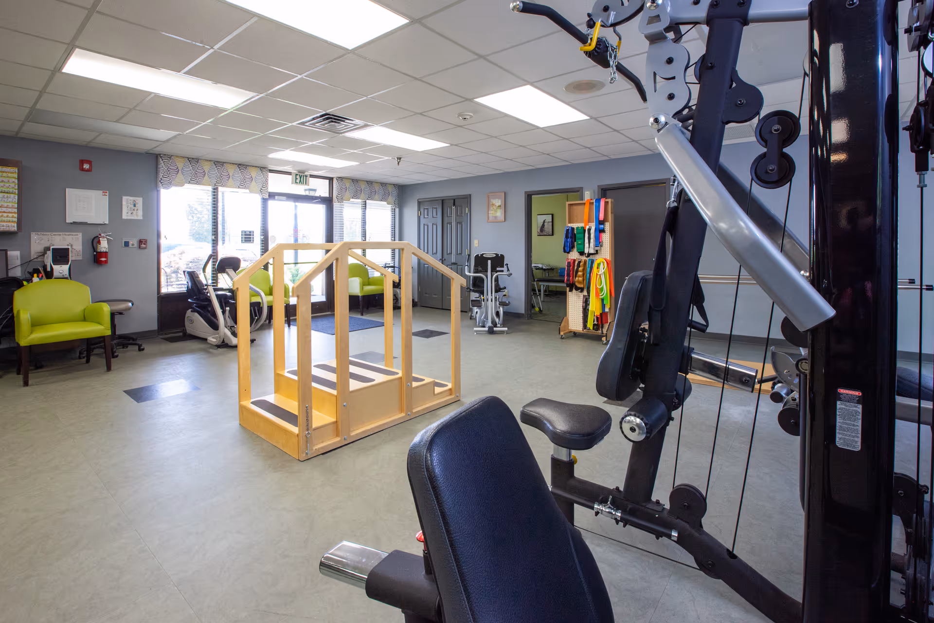 A bright therapy and fitness room with exercise equipment, parallel walking bars, and seating.