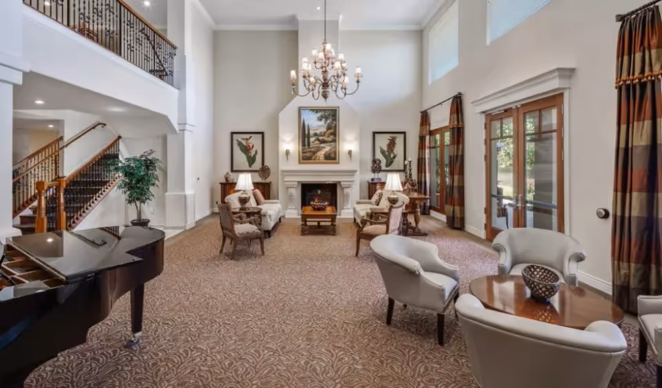 Spacious high-ceilinged common lounge with a chandelier, fireplace, grand piano, arranged seating, staircase, and glass doors to the outside.
