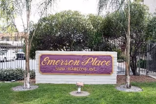 A purple Emerson Place sign mounted on a low stone wall in a landscaped front area with trees and a gated driveway behind it.