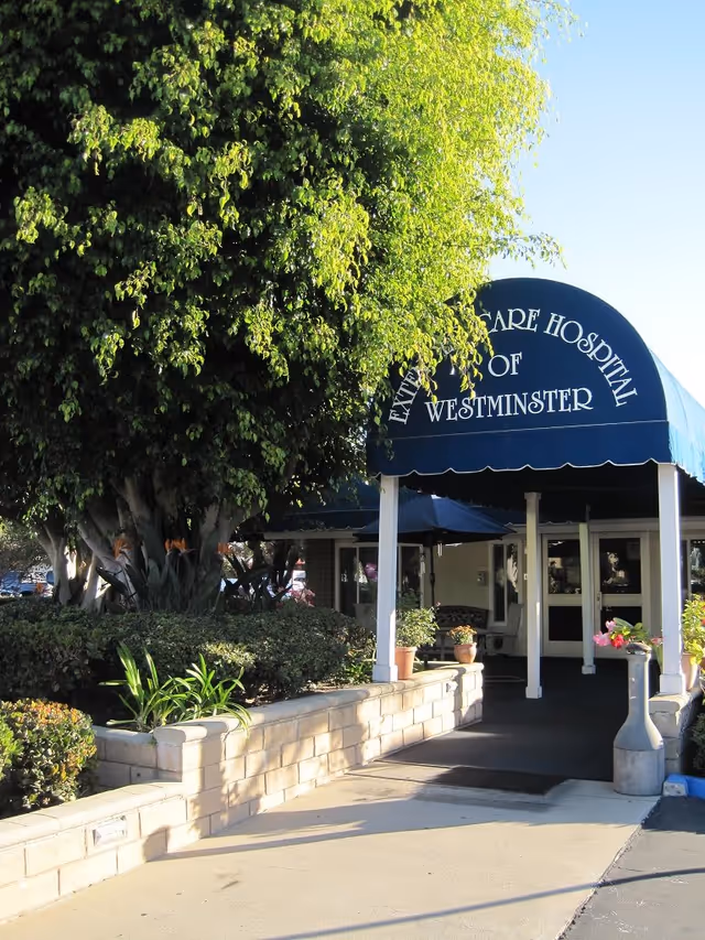 Entryway to Extended Care Hospital of Westminster with a blue awning, walkway, and surrounding landscaping.