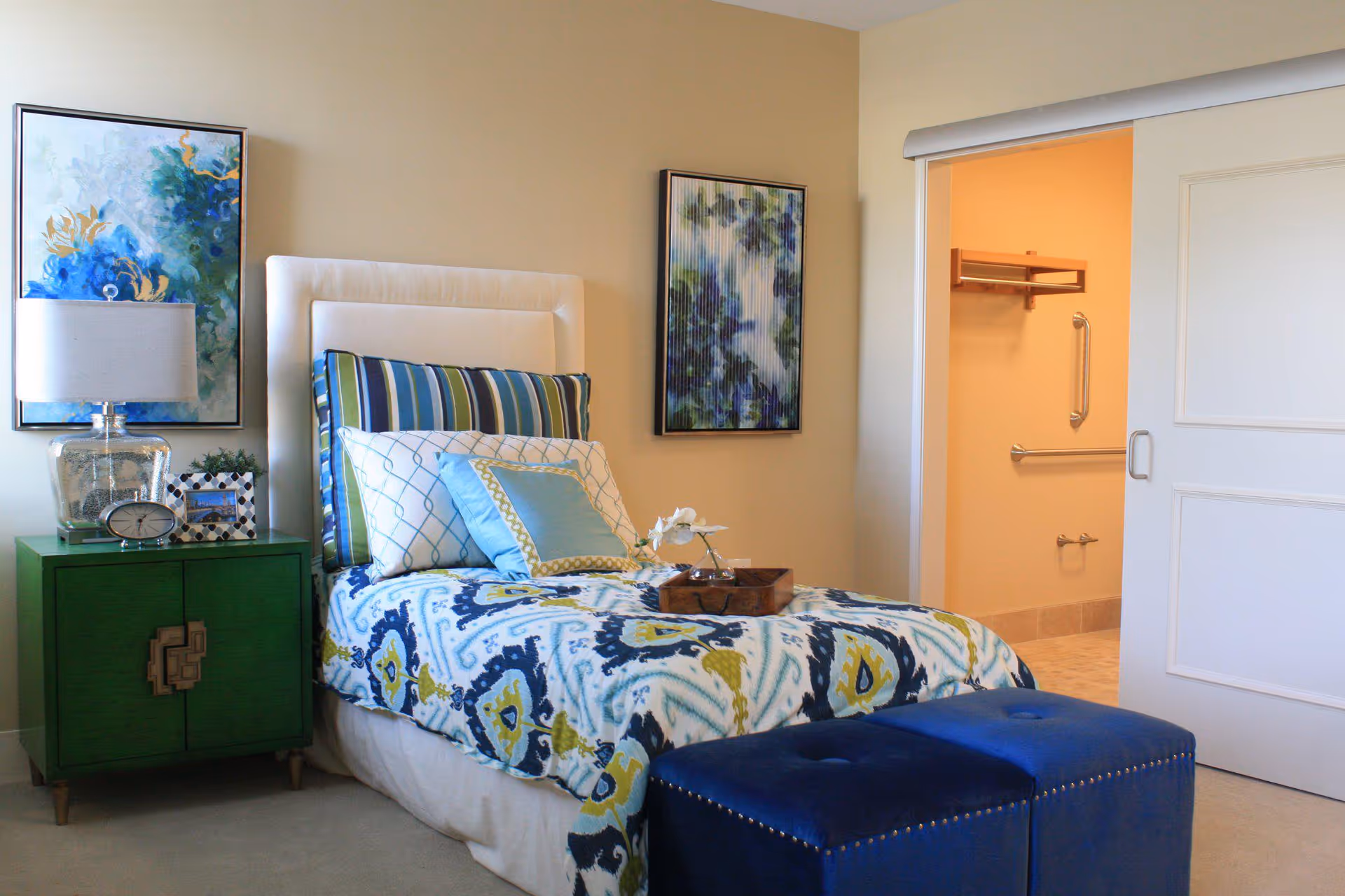 A neatly made single bed with a white upholstered headboard, colorful patterned bedding, and multiple pillows in a bedroom. Next to the bed is a green nightstand with a clear glass lamp, a clock, and a photo frame. Two abstract paintings hang on the beige walls. A sliding door is open, revealing a bathroom with grab bars and a tiled floor. Two blue ottomans are placed at the foot of the bed.