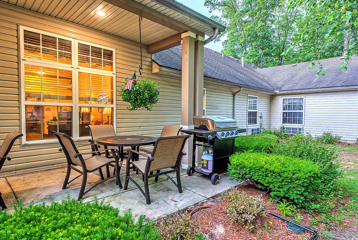 Outdoor patio area at Trail Creek Place Assisted Living featuring a round metal table with four chairs, a hanging flower pot with an American flag, a gas grill, and surrounding greenery and bushes. The building exterior has beige siding and multiple windows with warm interior lighting visible.