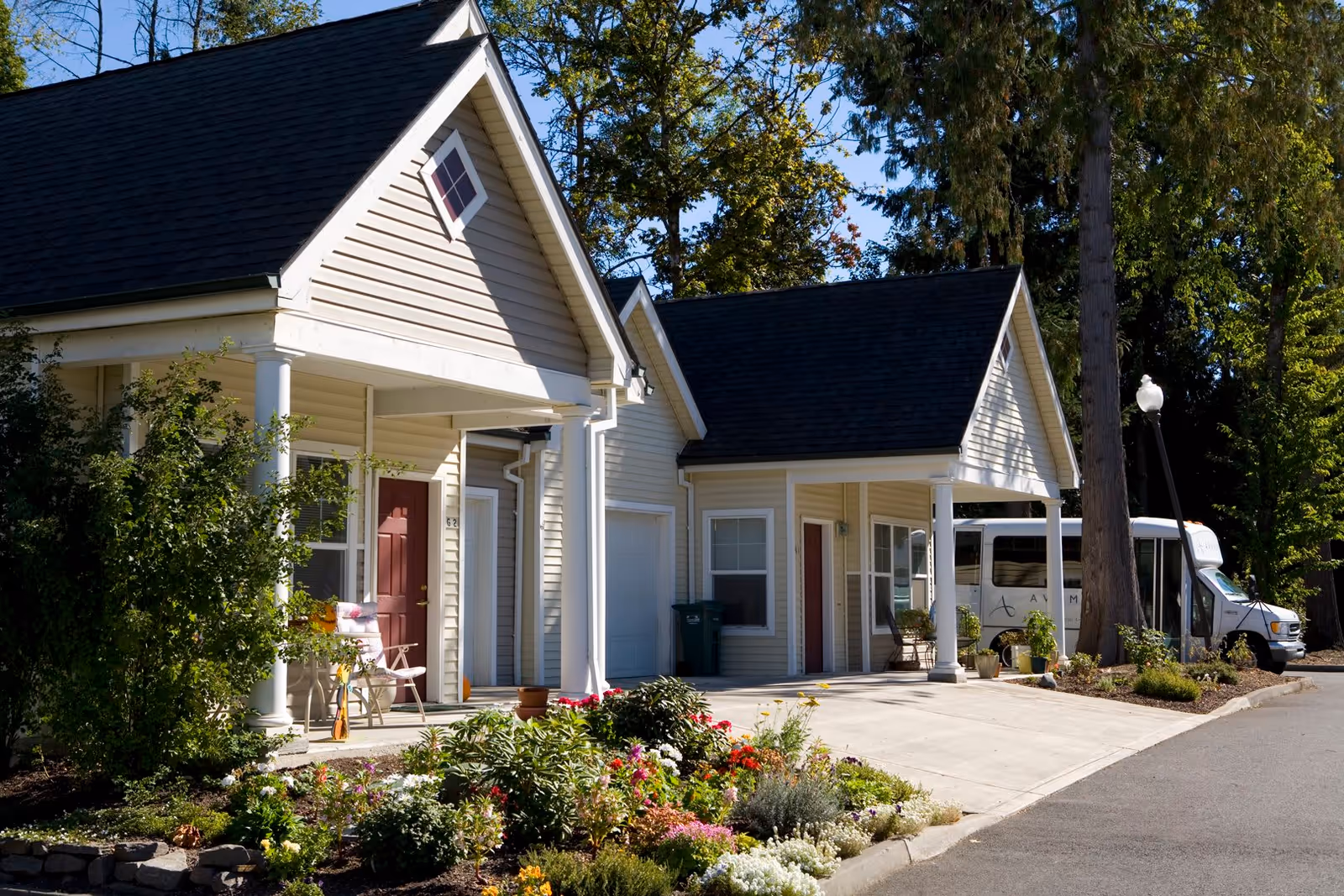 Exterior view of a senior living facility with beige siding and dark shingled roofs. The building features two small porches with white columns, potted plants, and a garden with colorful flowers in front. A white shuttle bus is parked on the driveway near the building, surrounded by tall trees and greenery.