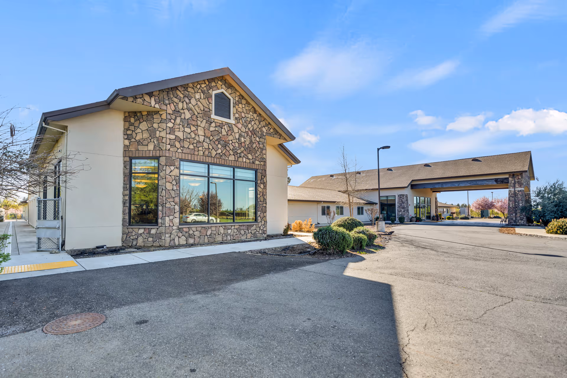 Exterior view of a senior living facility building with stone and beige walls, large windows, and a covered entrance driveway under a clear blue sky.
