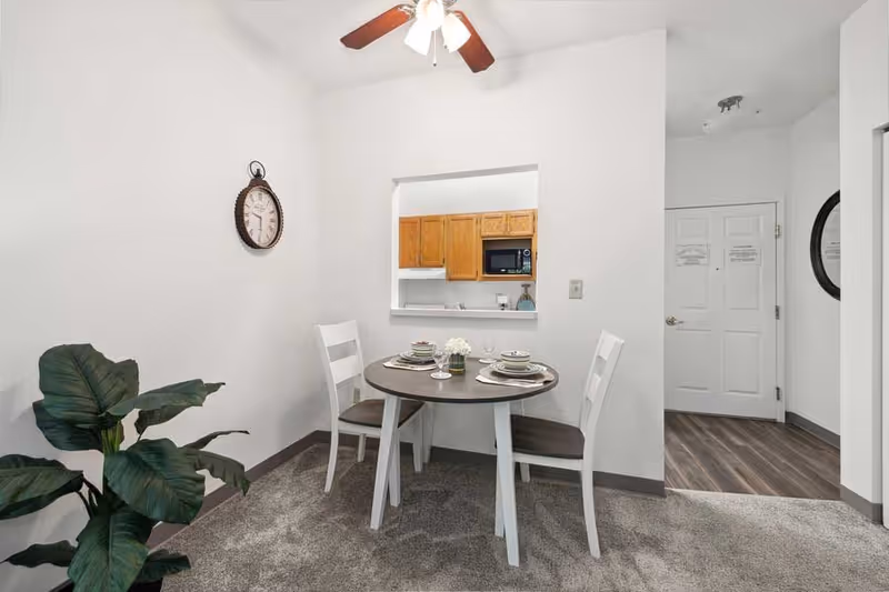 A small dining area with a round table set for two, featuring plates, bowls, and glasses. Two white chairs with dark seats are positioned around the table. A large green plant is placed on the left side near the wall. A wall clock hangs above the plant. The dining area opens to a kitchen with wooden cabinets and a microwave. The entrance door and a round mirror are visible in the background.