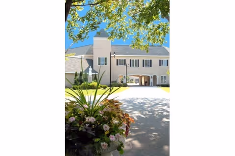 Front exterior of a beige two-story building with an arched entrance, driveway, and a flowering planter in the foreground beneath tree branches.