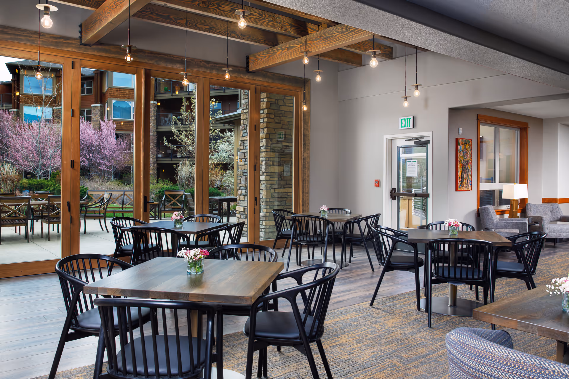Bright communal dining area with wooden tables and black chairs, small vases of flowers, and large glass doors opening to an outdoor patio and landscaped courtyard.