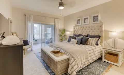 A spacious and well-lit bedroom featuring a large bed with a tufted headboard, multiple pillows, and a cozy blanket. The room includes a bench at the foot of the bed, a nightstand with a lamp, a dresser, a chair, and a sliding glass door leading to an outdoor patio. The walls are decorated with framed artwork, and the room has a neutral color palette with soft lighting.