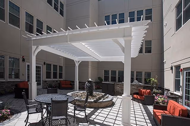 Outdoor courtyard area with a white pergola providing partial shade over a round table with chairs. Surrounding the area are cushioned seating arrangements with red cushions, potted plants, and windows of the building enclosing the space.
