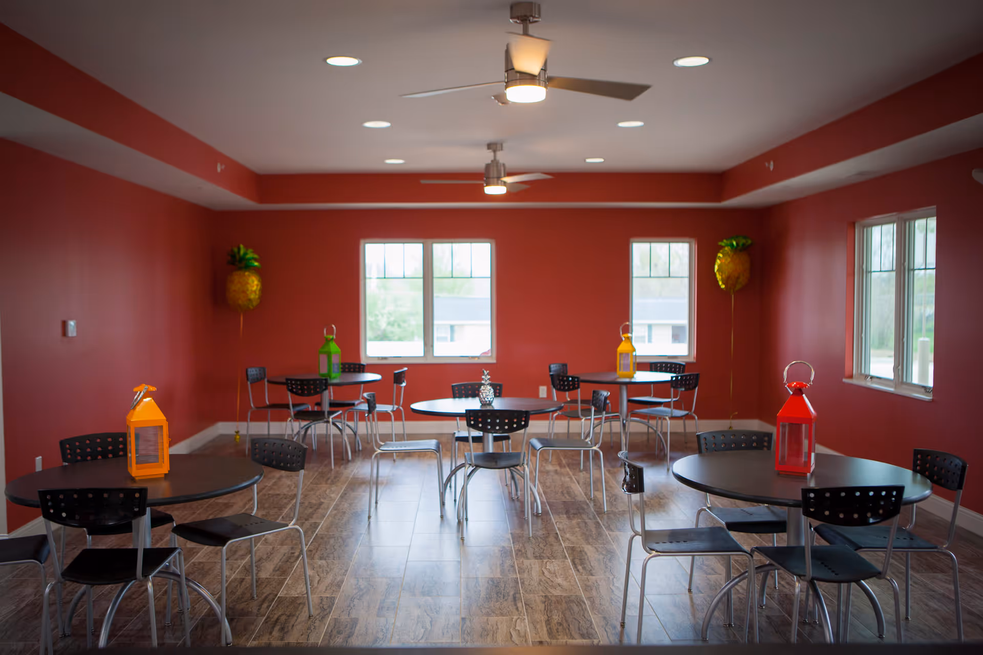 Bright common room with red walls, round tables and black chairs on a wood-look floor beneath ceiling fans and windows.