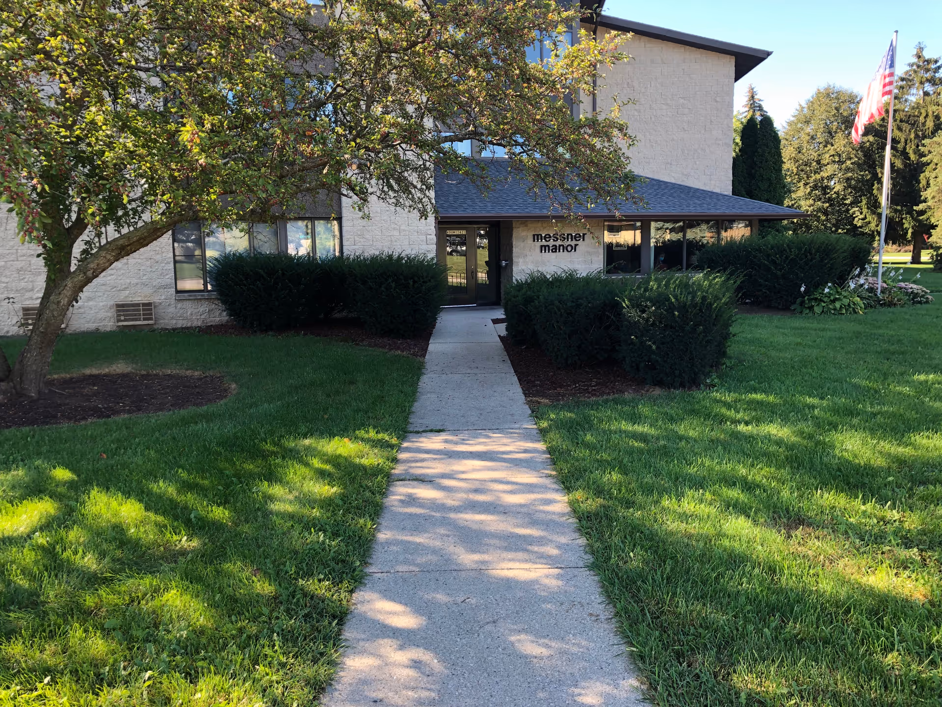 Sidewalk leads through a green lawn and trees to the front entrance of a low-rise building with bushes and a sign reading "messner manor".
