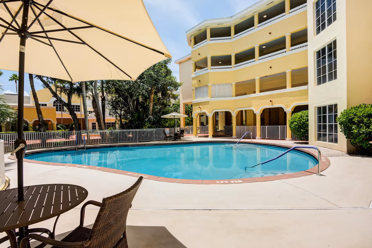 Outdoor swimming pool area at The Preserve at Palm-Aire with a round table and chairs under a large beige umbrella in the foreground, surrounded by a yellow multi-story building and lush greenery under a clear blue sky.