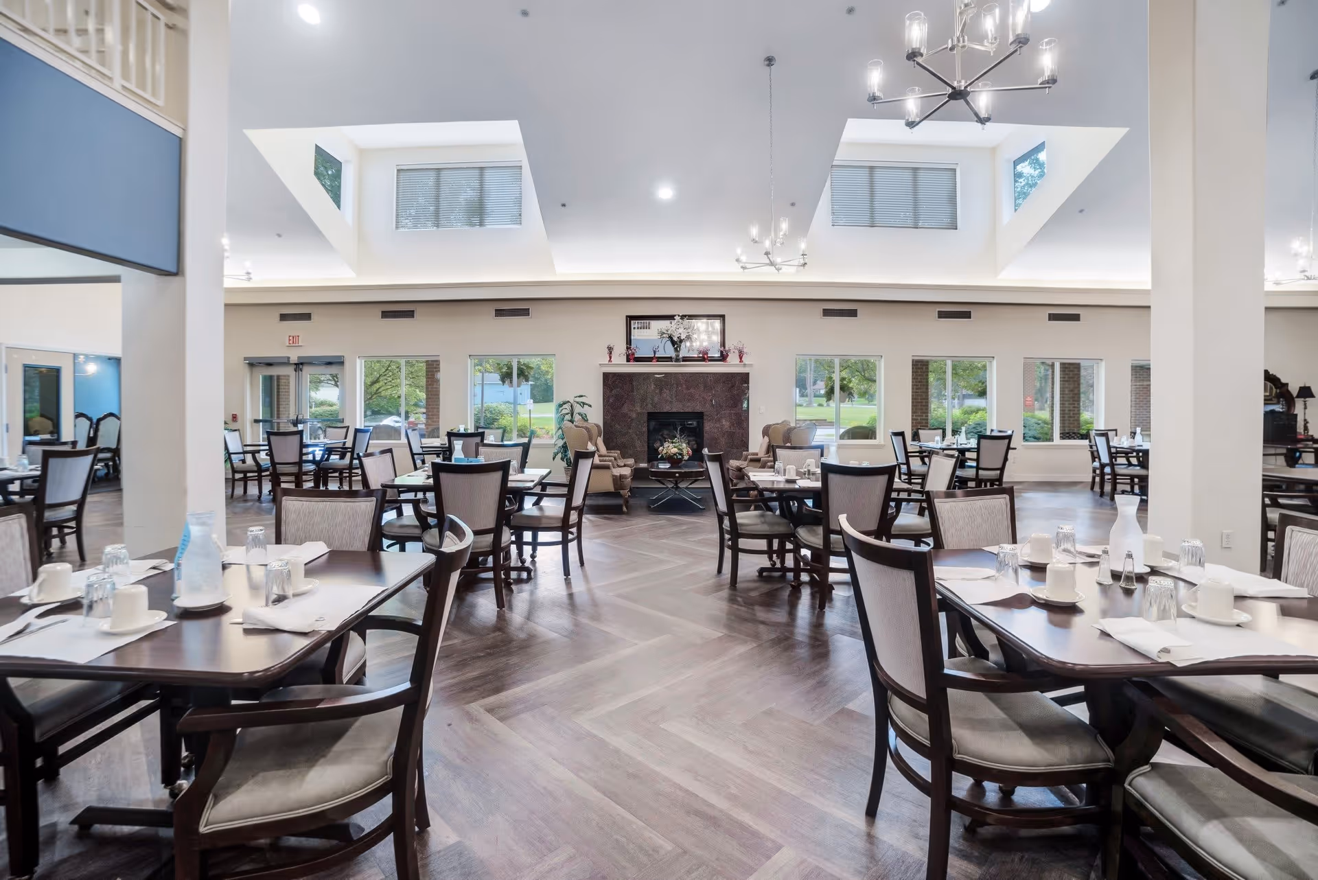 A spacious dining room with multiple dark wood tables and chairs arranged neatly. Each table is set with white cups, glasses, napkins, and silverware. The room has large windows letting in natural light, a fireplace in the center of the far wall, and modern chandeliers hanging from the ceiling with skylights above.