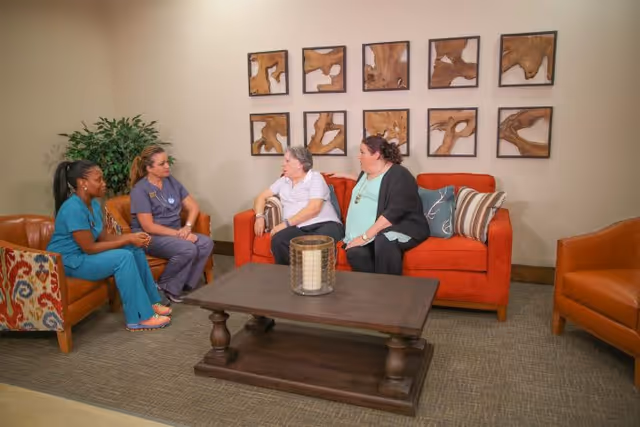 Two healthcare workers in scrubs sitting on a brown leather chair and an orange sofa, talking with two women seated on an orange sofa in a cozy living room setting with a wooden coffee table and abstract wooden wall art.