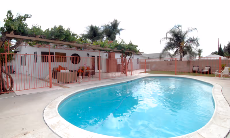 Outdoor swimming pool with clear blue water surrounded by a concrete deck. Behind the pool is a fenced patio area with outdoor seating, a pergola with greenery, and palm trees in the background.